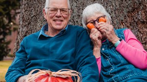 elderly couple leaning on a tree trunk holding colourful pumpkins